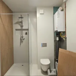 Nowoczesna łazienka w trakcie wykańczania: kabina prysznicowa typu walk-in z deszczownicą, wisząca toaleta, piec gazowy na ścianie, białe i drewnopodobne płytki, wystające przewody elektryczne...