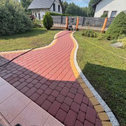 Winclear mycie okien i elewacji - Czerwona kostka brukowa z żółto-szarym obrzeżem wijąca się przez zielony trawnik w kierunku domu i ogrodzenia; widoczny wąż ciśnieniowy i myjka ciśnieniowa w tle.