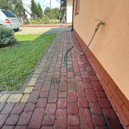 Winclear mycie okien i elewacji - Czerwona kostka brukowa przed budynkiem z kranem i wężem ogrodowym, widoczne ślady wilgoci sugerujące niedawne mycie.