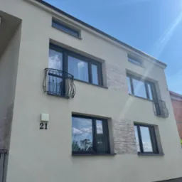 Nowoczesny budynek dwupiętrowy z jasnobeżową elewacją, ciemnymi ramami okiennymi i małymi balkonami z kutą balustradą, numer domu '21' widoczny na elewacji, fragment dekoracyjnej okładziny...