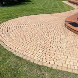 Winclear mycie okien i elewacji - Kremowa kostka brukowa ułożona w łukowaty wzór, obrzeża z okrągłych kostek, trawnik, panele słoneczne w tle, Jasionka. Czysta nawierzchnia.