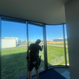 Winclear mycie okien i elewacji - Pracownik w czarnym stroju myje okno w biurowcu za pomocą ściągaczki. Widok na zewnątrz ukazuje budynki i zielony teren. Okno o nietypowym, zakrzywionym kształcie.