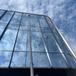 Winclear mycie okien i elewacji - Mycie szklanej elewacji nowoczesnego budynku z użyciem wysięgnika i szczotki. Widoczne krople wody spływające po oknach i odbicie nieba w czystych szybach.