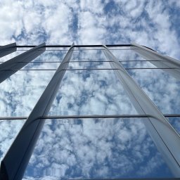 Winclear mycie okien i elewacji - Szklana fasada budynku biurowego, odbijająca błękitne niebo z chmurami. Widok z dołu, pionowe linie podziału okien.