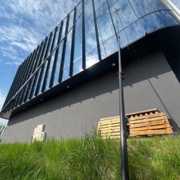 Winclear mycie okien i elewacji - Nowoczesny budynek z lustrzanymi oknami mytymi teleskopową lancą. W tle błękitne niebo z chmurami. Na dole trawa i palety.