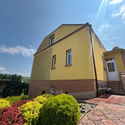 Winclear mycie okien i elewacji - Żółta elewacja domu z brązowym cokołem, widok od dołu z ogrodem i kostką brukową. Słońce odbija się od okien. Widoczna ławka.