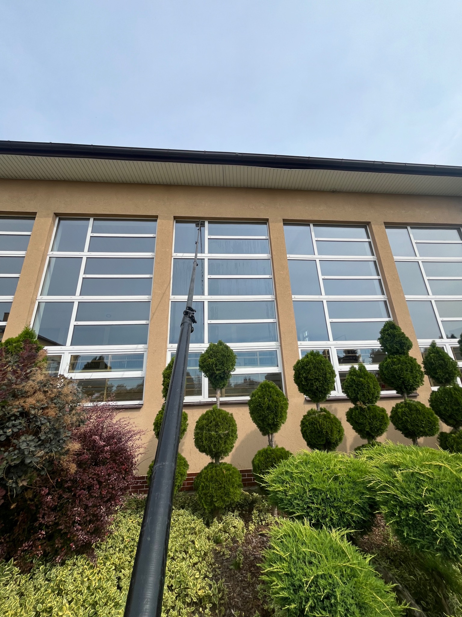 Czarny teleskopowy kij do mycia okien sięga do wysokich okien budynku z beżową elewacją, otoczonego ozdobnymi krzewami i topiarami.