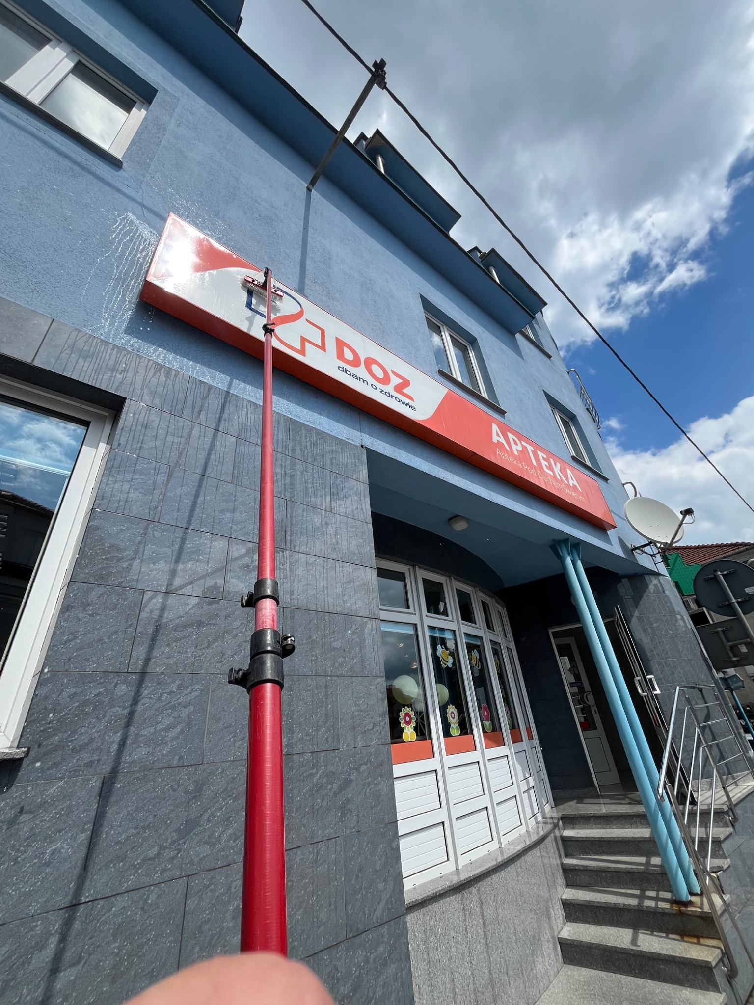 Mycie elewacji budynku apteki za pomocą teleskopowej lancy z szczotką, widać spływającą wodę. Niebieska elewacja i szyld reklamowy.