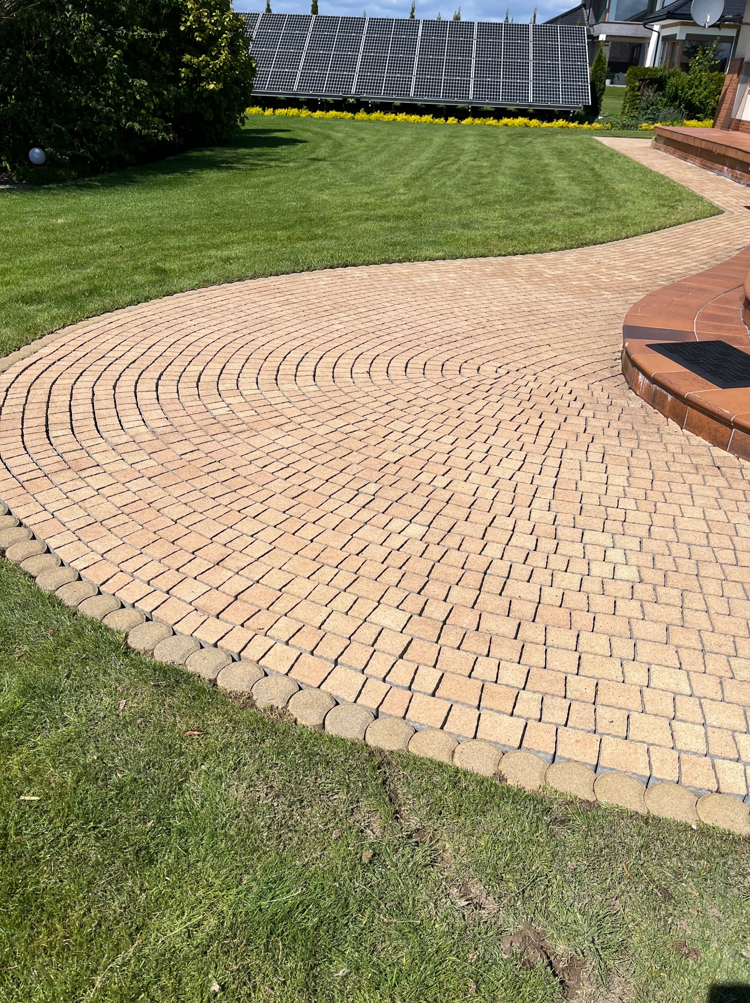 Kremowa kostka brukowa ułożona w łukowaty wzór, obrzeża z okrągłych kostek, trawnik, panele słoneczne w tle, Jasionka. Czysta nawierzchnia.