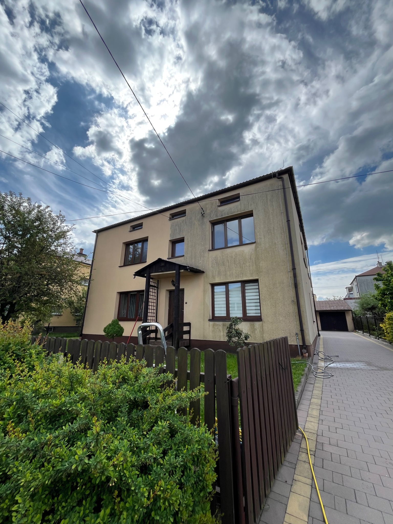 Dwukondygnacyjny dom z beżową elewacją, widoczny z poziomu gruntu. Na elewacji widoczne zabrudzenia. W tle błękitne niebo z chmurami i fragment chodnika z kostki.