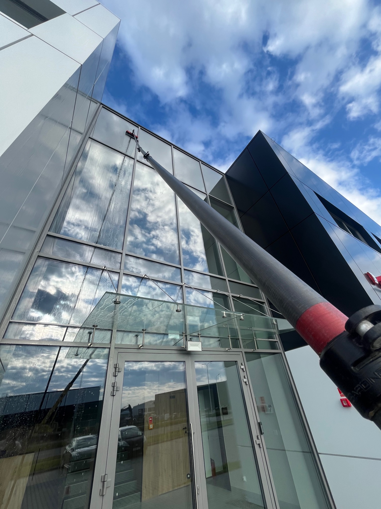 Mycie szklanej elewacji biurowca z użyciem teleskopowej lancy i szczotki, z widocznymi smugami wody. W tle błękitne niebo z chmurami odbijające się w szybach.