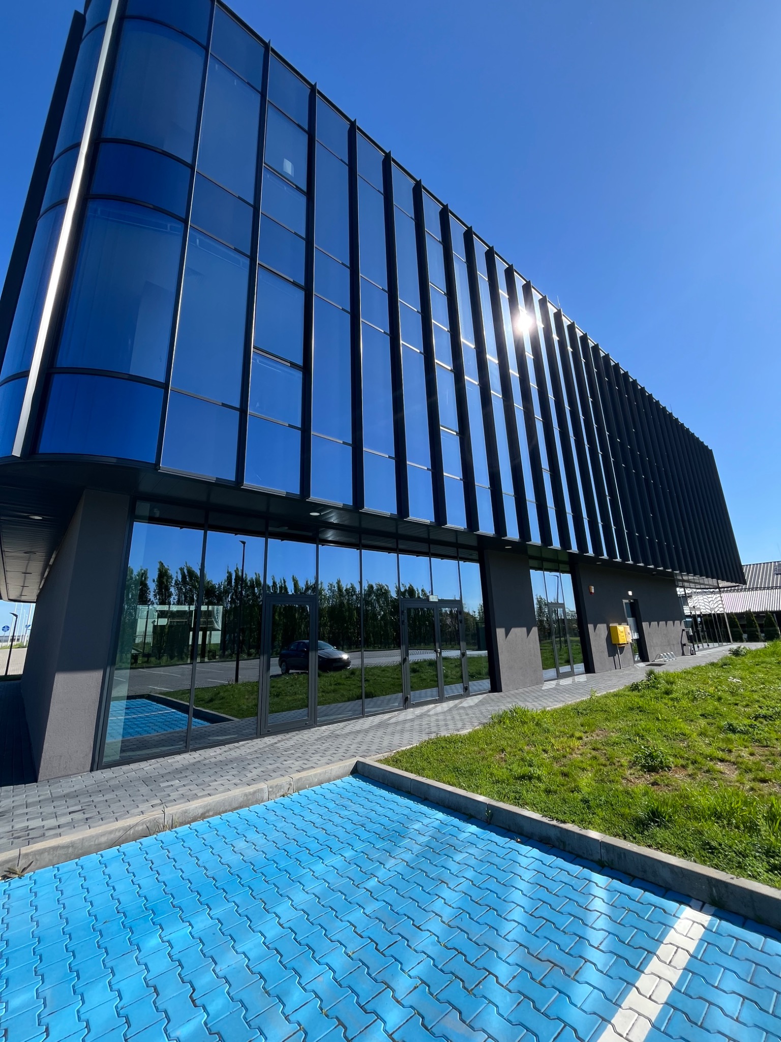 Nowoczesny biurowiec z lustrzanymi szybami odbijającymi niebo i zieleń, z niebieską kostką brukową na parkingu. Słońce odbija się w oknach.