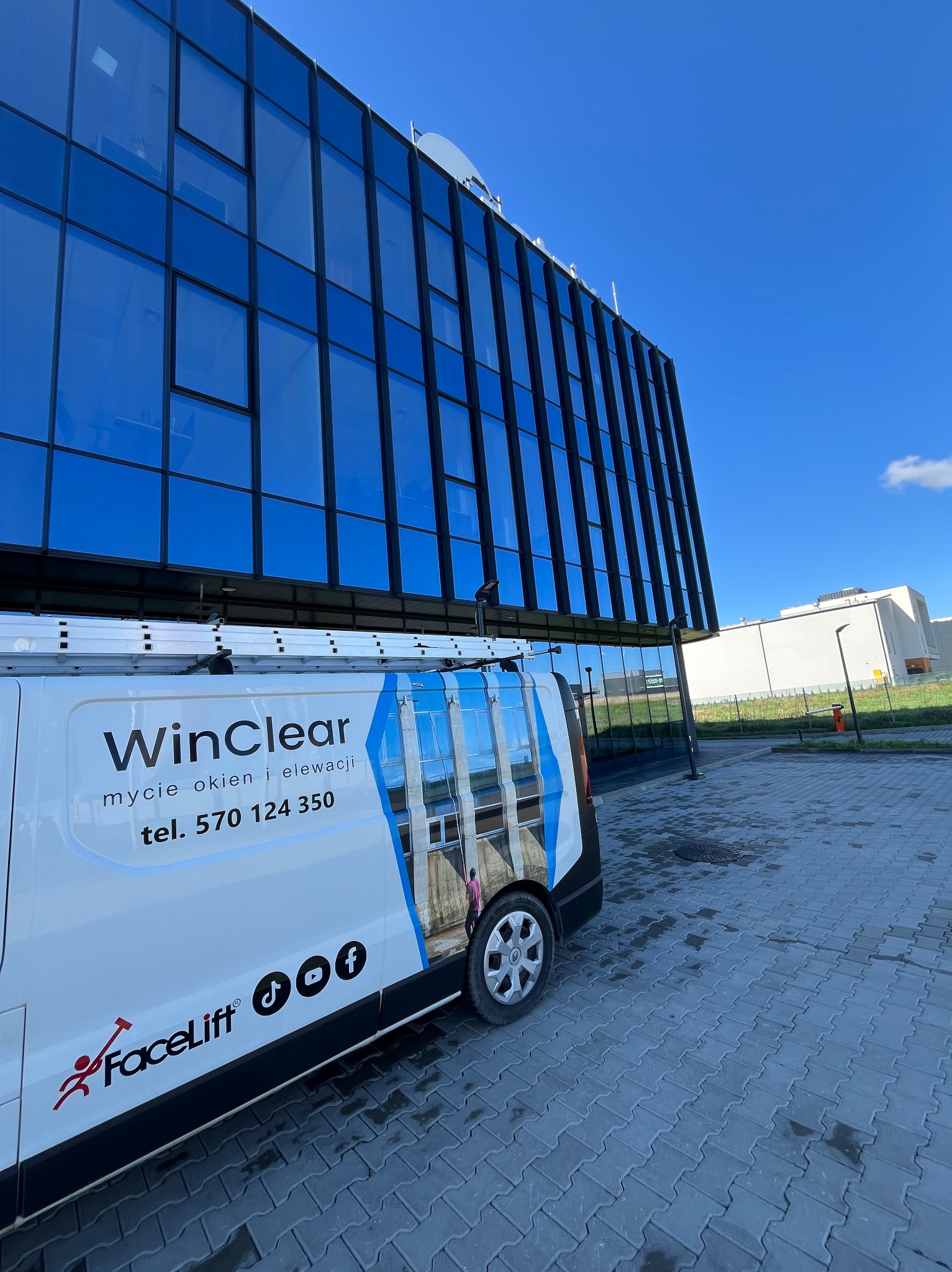 Biały bus z logo WinClear i numerem telefonu na tle nowoczesnego biurowca z niebieskimi oknami. Usługi mycia okien i elewacji. Błękitne niebo w tle.