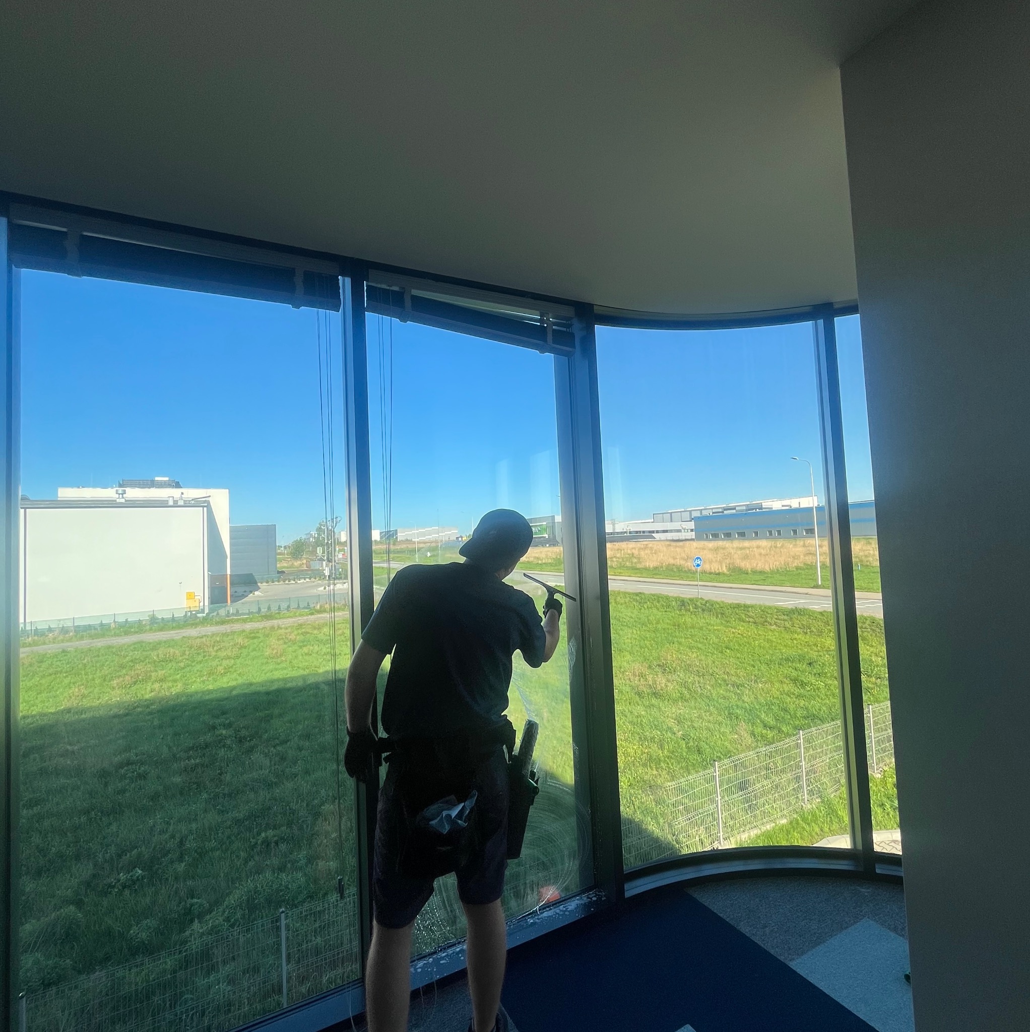 Pracownik w czarnym stroju myje okno w biurowcu za pomocą ściągaczki. Widok na zewnątrz ukazuje budynki i zielony teren. Okno o nietypowym, zakrzywionym kształcie.