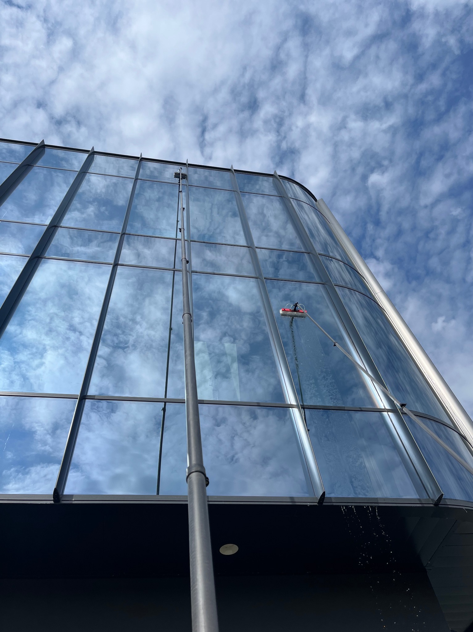 Mycie szklanej elewacji nowoczesnego budynku z użyciem wysięgnika i szczotki. Widoczne krople wody spływające po oknach i odbicie nieba w czystych szybach.