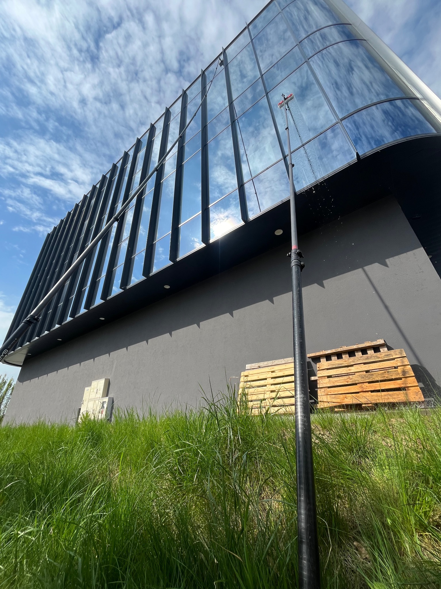 Nowoczesny budynek z lustrzanymi oknami mytymi teleskopową lancą. W tle błękitne niebo z chmurami. Na dole trawa i palety.