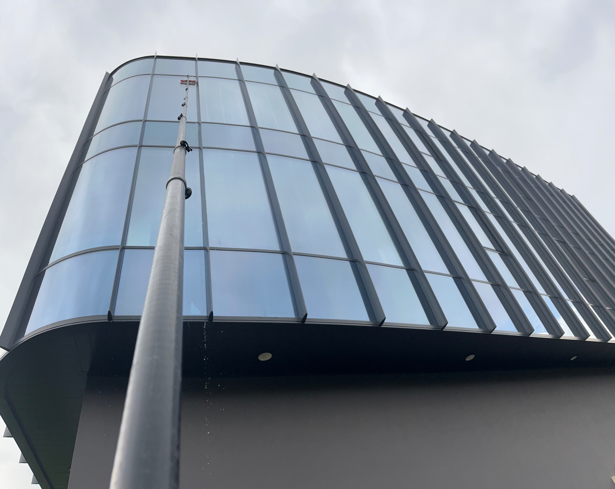 Mycie szklanej elewacji budynku biurowego za pomocą teleskopowej lancy. Widoczne ślady wody spływającej po szybach, pochmurne niebo w tle.