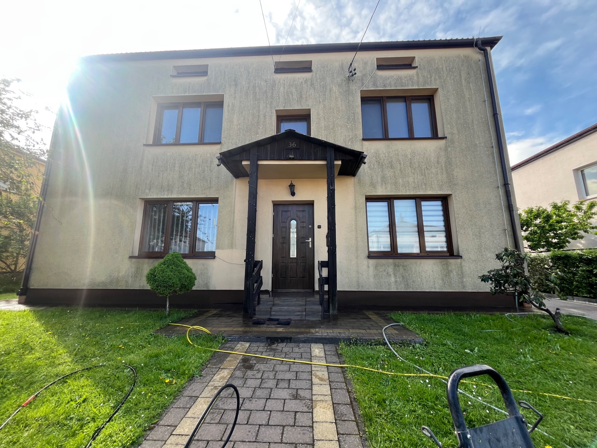 Dwupiętrowy dom z beżową elewacją po umyciu, widoczny fragment kostki brukowej po czyszczeniu, węże i sprzęt do mycia. Słoneczny dzień.