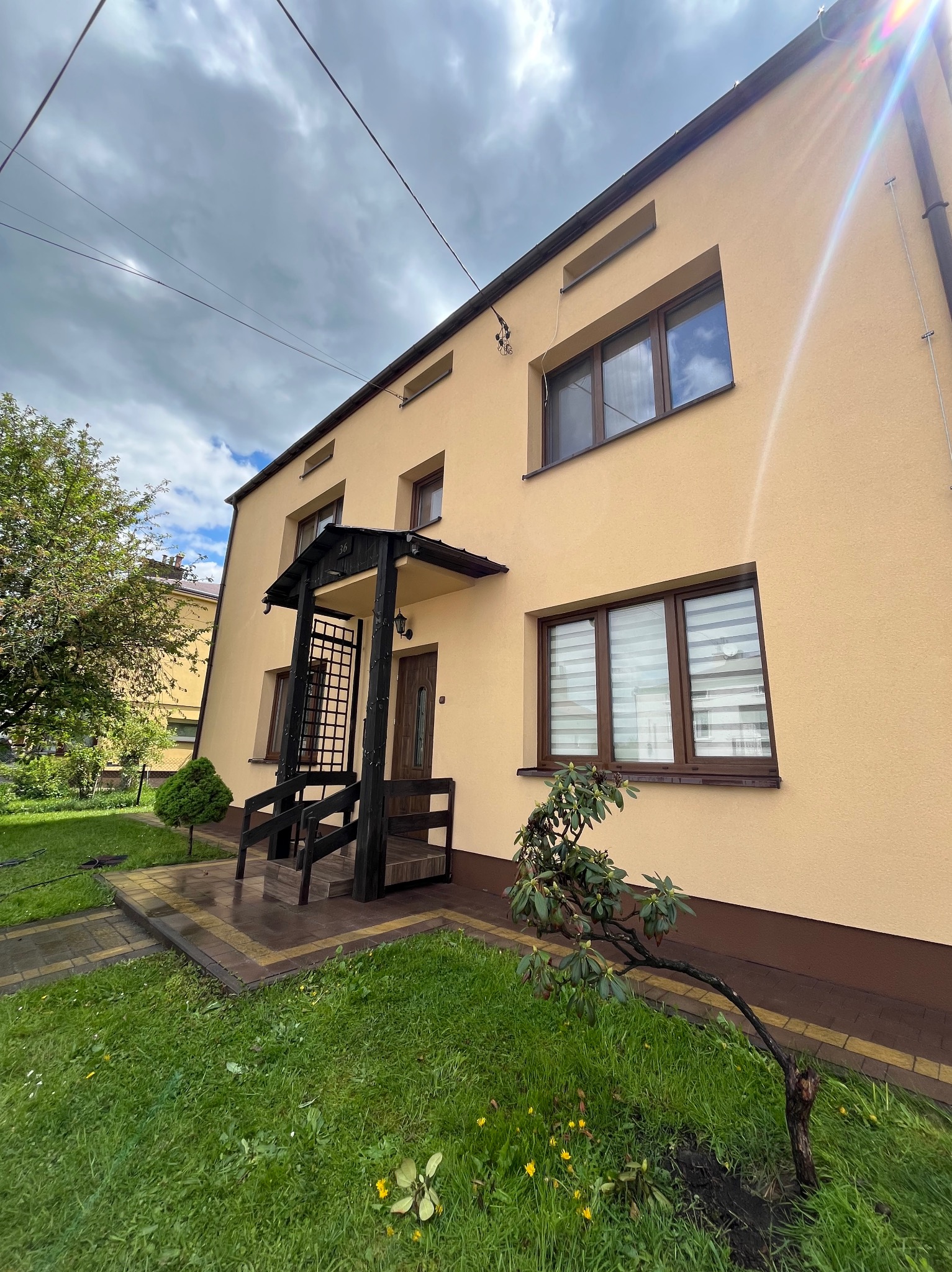 Dwukondygnacyjny dom z beżową elewacją, ciemnymi oknami i zadaszonym wejściem. Widoczne elementy krajobrazu: trawnik, drzewa i krzewy.