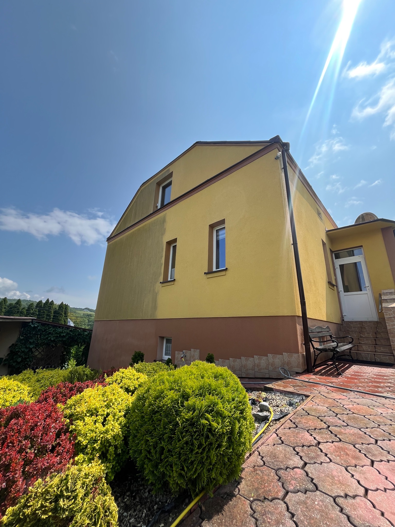 Żółta elewacja domu po czyszczeniu, z widoczną różnicą w kolorze. Czerwona kostka brukowa i dekoracyjny ogród. Jasne, słoneczne ujęcie.