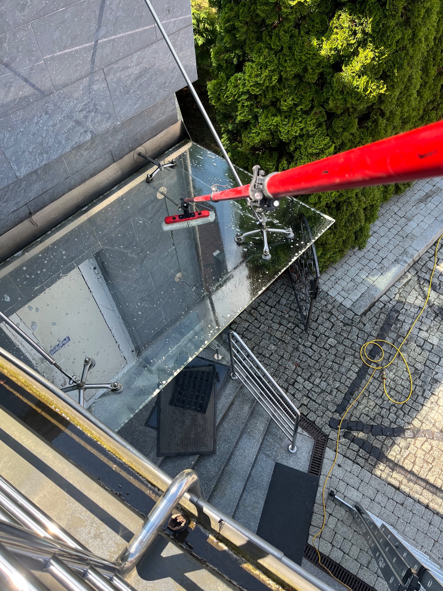 Czerwony kij z szczotką myje szklany daszek nad wejściem do budynku. Widoczna kostka brukowa i wąż ogrodowy. Ujęcie z góry.