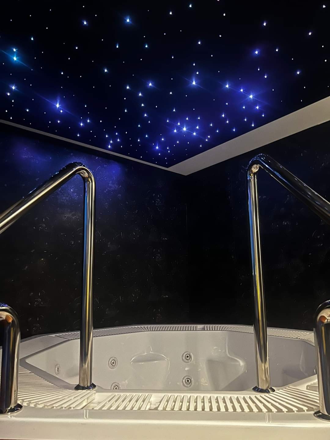 Wanna z hydromasażem w pomieszczeniu z ciemnymi ścianami i sufitem imitującym rozgwieżdżone niebo, z chromowanymi poręczami ułatwiającymi wejście.