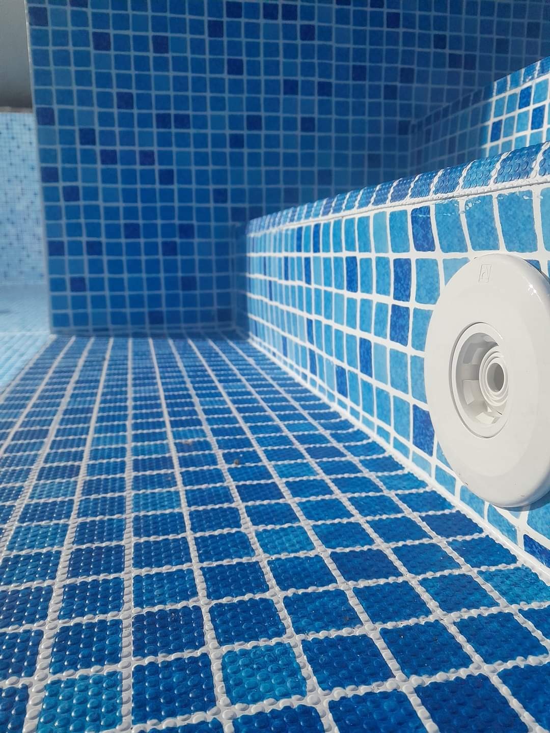 Wnętrze basenu wyłożonego mozaiką w odcieniach błękitu i bieli, z widocznym dyszą wlotową wody, oświetlone naturalnym światłem.