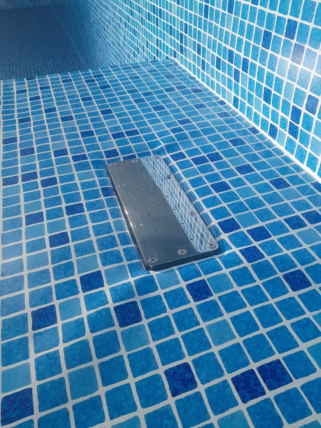 Wykończone wnętrze niebieskiego basenu mozaiką z zamontowanym, metalowym odpływem dennym z widocznymi śrubami.