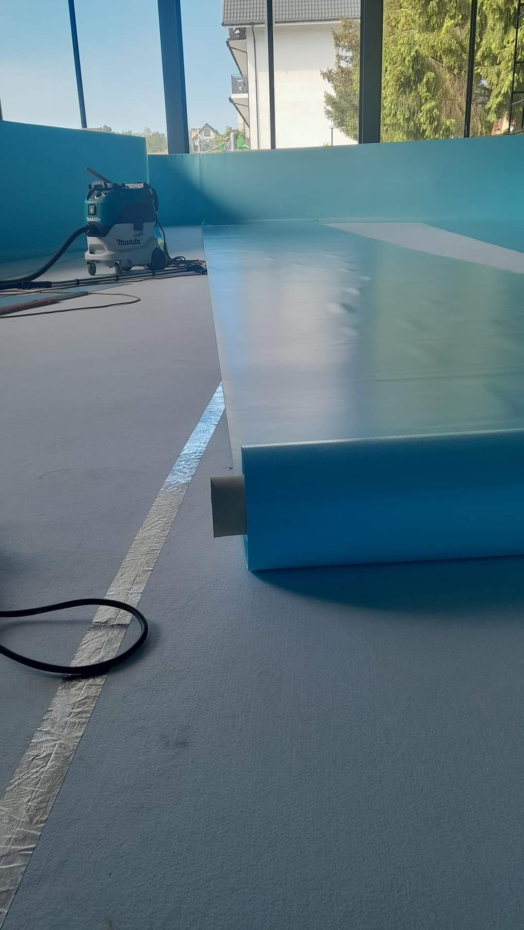 Montaż niebieskiej folii izolacyjnej w nowym basenie krytym, widoczny odkurzacz przemysłowy Makita, taśma uszczelniająca na podłodze i okna z widokiem na budynek mieszkalny.
