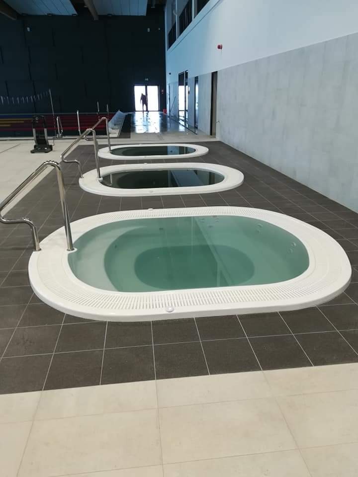 Trzy owalne jacuzzi z białym obramowaniem, wypełnione wodą, ustawione w rzędzie na ciemnoszarej podłodze w hali basenowej, z metalowymi poręczami ułatwiającymi wejście.