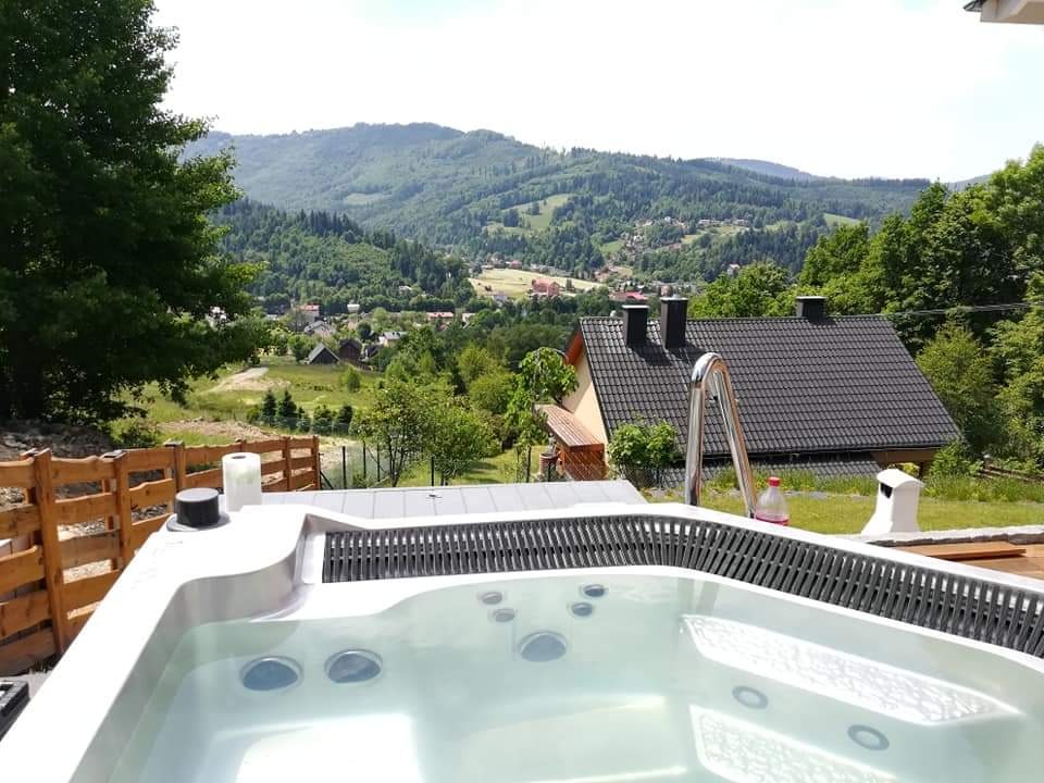 Biały basen jacuzzi z metalową drabinką na tle górskiego krajobrazu i zabudowań mieszkalnych.