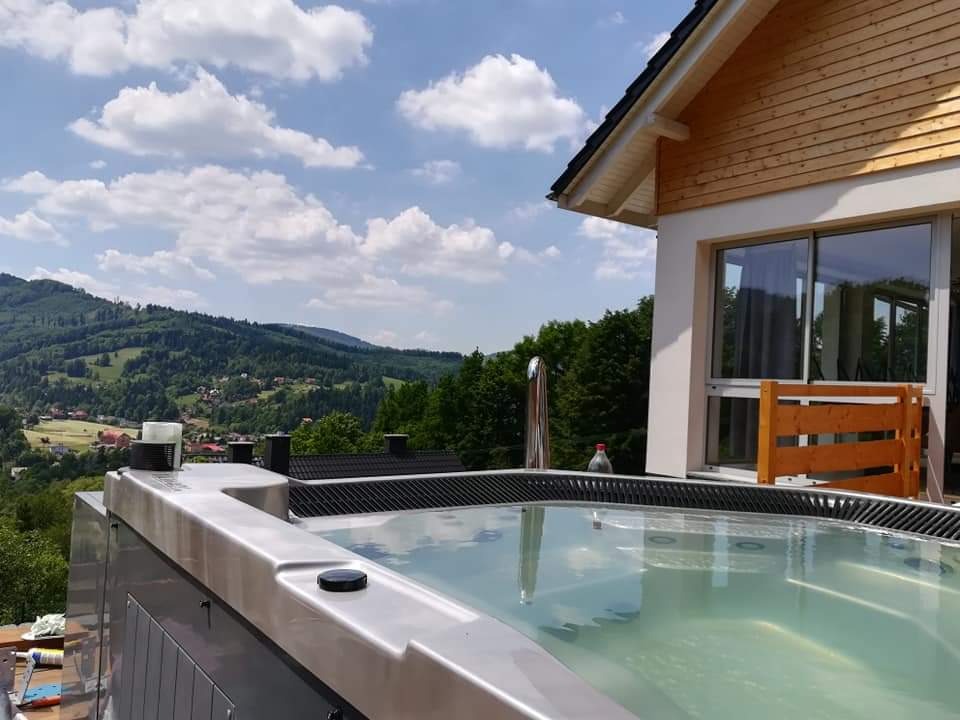 Zewnętrzne jacuzzi ze stali nierdzewnej na tarasie z widokiem na górzysty krajobraz, obok budynku z drewnianą elewacją i dużym oknem.