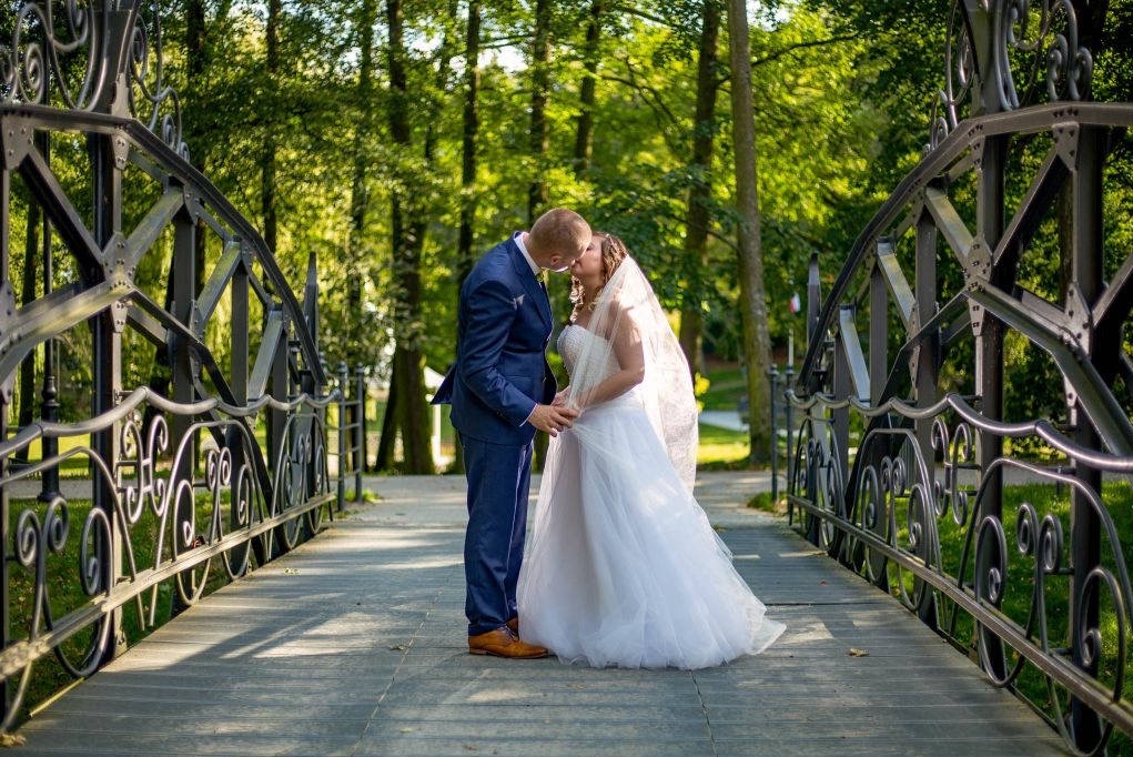 Para młoda całująca się na ozdobnym, metalowym moście w parku, z widocznymi drzewami w tle.