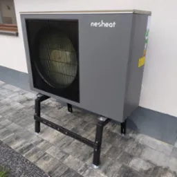 Szara jednostka zewnętrzna z logo 'nesheat' na czarnym metalowym stelażu, ustawiona na szarej kostce brukowej przy elewacji budynku z widocznym oknem i drzwiami.