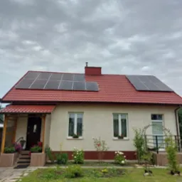 Dom jednorodzinny z czerwoną dachówką i panelami słonecznymi na dachu, widoczne kwiaty w oknach i roślinność w ogrodzie.