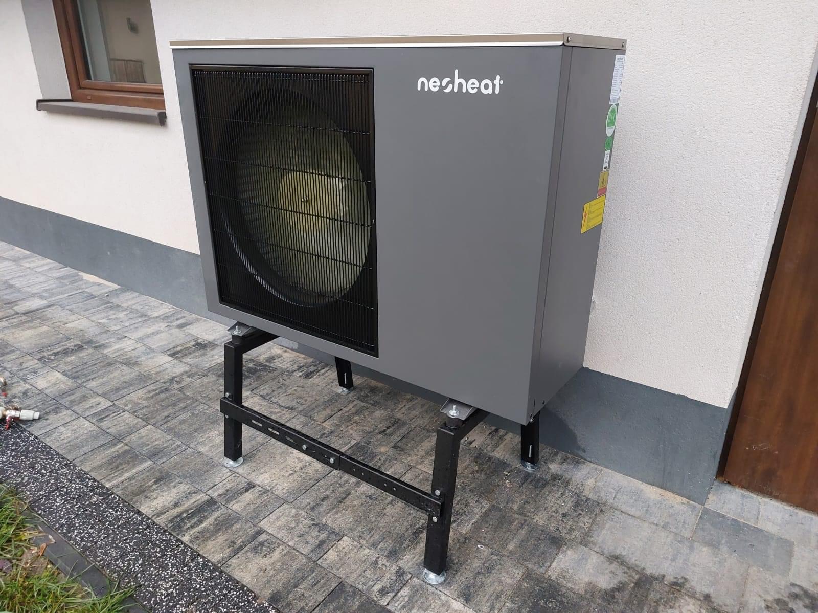 Szara jednostka zewnętrzna z logo 'nesheat' na czarnym metalowym stelażu, ustawiona na szarej kostce brukowej przy elewacji budynku z widocznym oknem i drzwiami.