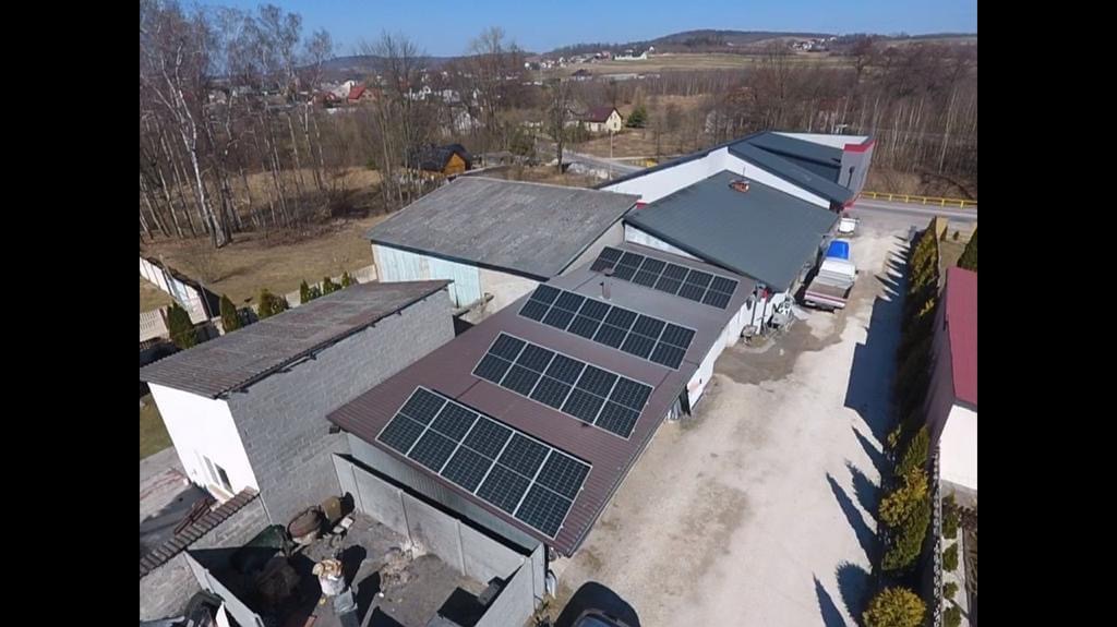 Widok z lotu ptaka na duży budynek z zainstalowanymi panelami słonecznymi na dachu, w tle zabudowania wiejskie i drzewa.