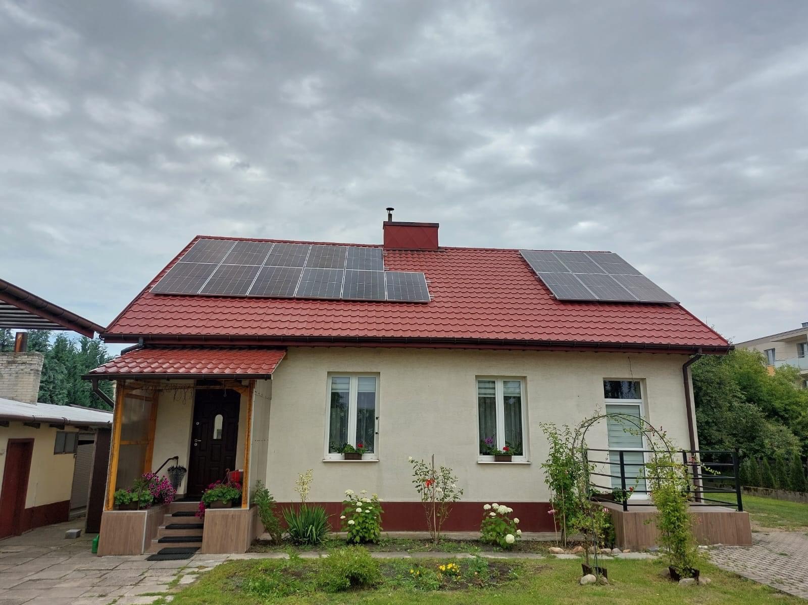 Dom jednorodzinny z czerwoną dachówką i panelami słonecznymi na dachu, widoczne kwiaty w oknach i roślinność w ogrodzie.