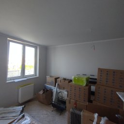 MatchIn Dominik Szyda - Wnętrze pokoju po malowaniu ścian na biało, widoczne kartony i elementy wyposażenia przygotowane do dalszych prac wykończeniowych. Duże okno z widokiem na zewnątrz.