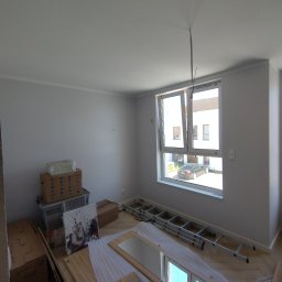 MatchIn Dominik Szyda - Wnętrze pokoju po malowaniu ścian na biało, z widoczną drabiną, kartonami i elementami wyposażenia. Okno otwarte, w tle budynek i samochód.