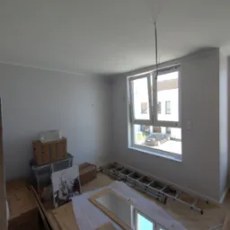 Wnętrze pokoju po malowaniu ścian na biało, z widoczną drabiną, kartonami i elementami wyposażenia. Okno otwarte, w tle budynek i samochód.