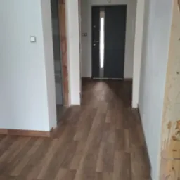 Ułożone panele podłogowe w korytarzu, widoczne fragmenty ścian w trakcie wykończenia i drzwi wejściowe w głębi. Podłoga w odcieniach brązu, ściany białe.