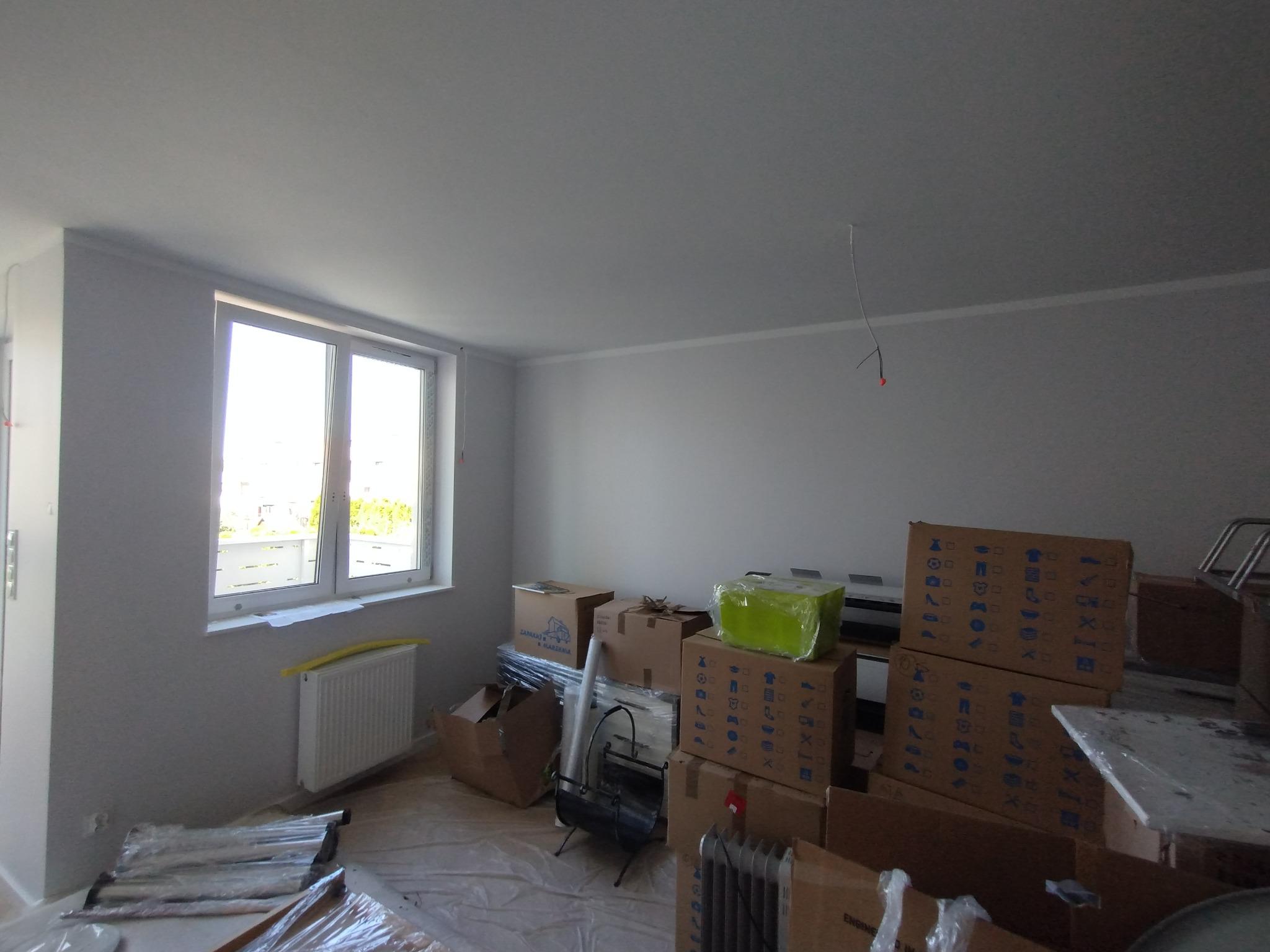 Wnętrze pokoju po malowaniu ścian na biało, widoczne kartony i elementy wyposażenia przygotowane do dalszych prac wykończeniowych. Duże okno z widokiem na zewnątrz.