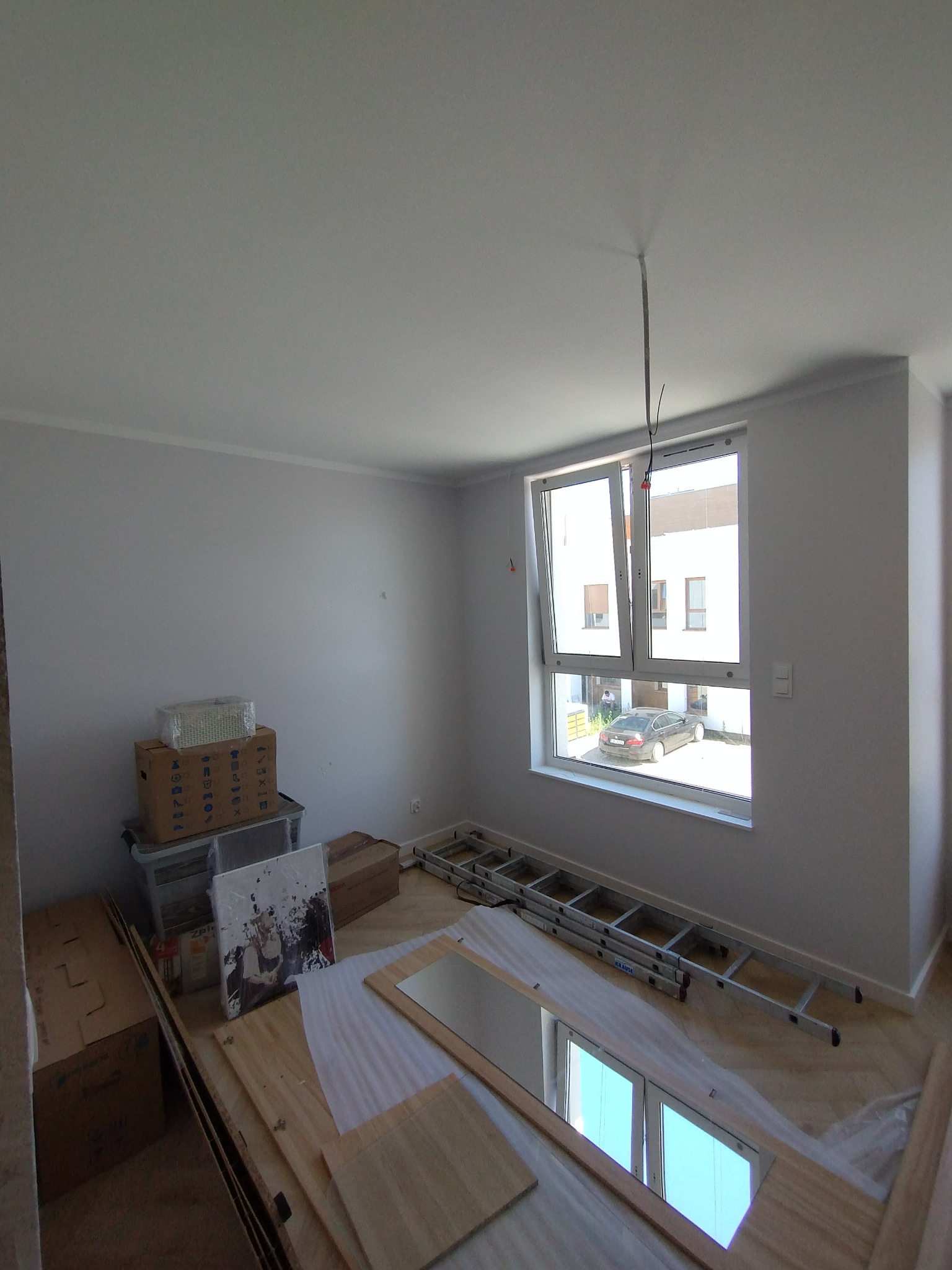 Wnętrze pokoju po malowaniu ścian na biało, z widoczną drabiną, kartonami i elementami wyposażenia. Okno otwarte, w tle budynek i samochód.