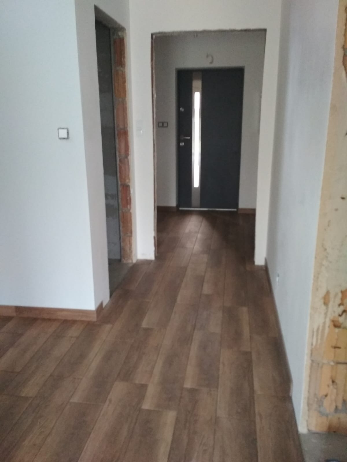 Ułożone panele podłogowe w korytarzu, widoczne fragmenty ścian w trakcie wykończenia i drzwi wejściowe w głębi. Podłoga w odcieniach brązu, ściany białe.