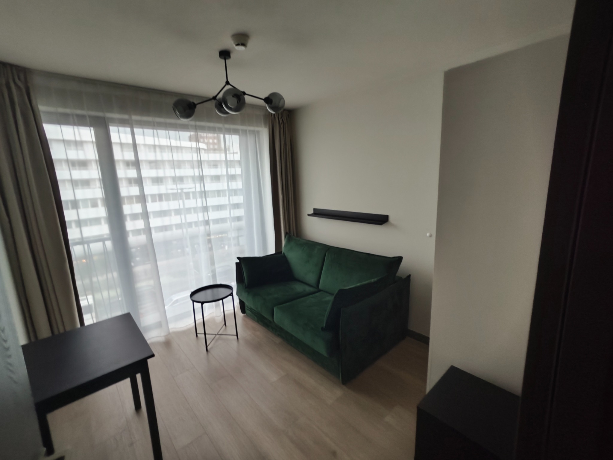 Jasne wnętrze po remoncie z zieloną sofą i drewnianą podłogą. Nowoczesna lampa sufitowa i minimalistyczne meble. Duże okno z widokiem na miasto.