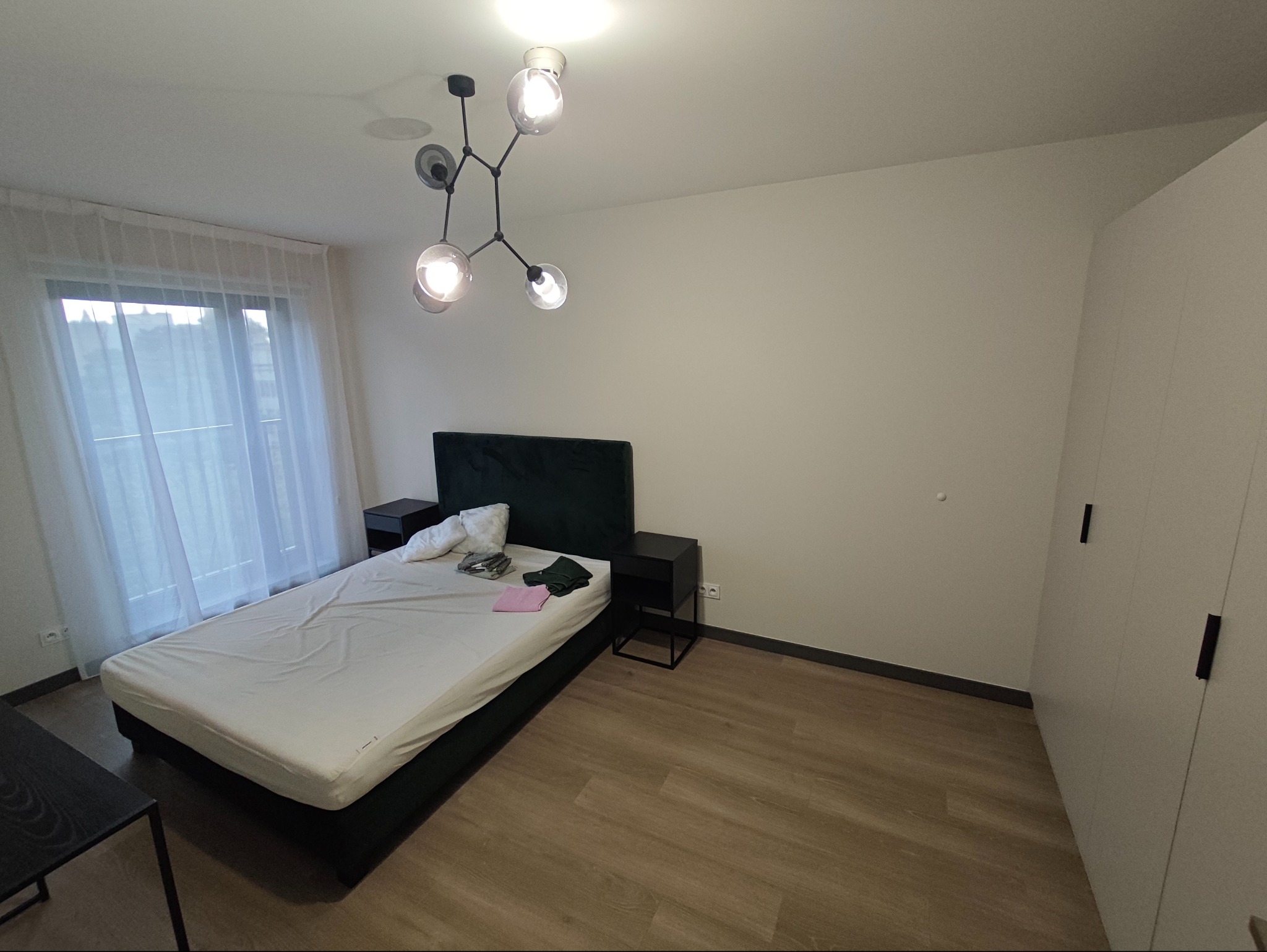 Jasna sypialnia z łóżkiem z zielonym wezgłowiem, drewnianą podłogą i nowoczesną lampą sufitową. Widok na okno z białymi zasłonami i minimalistyczną szafę.