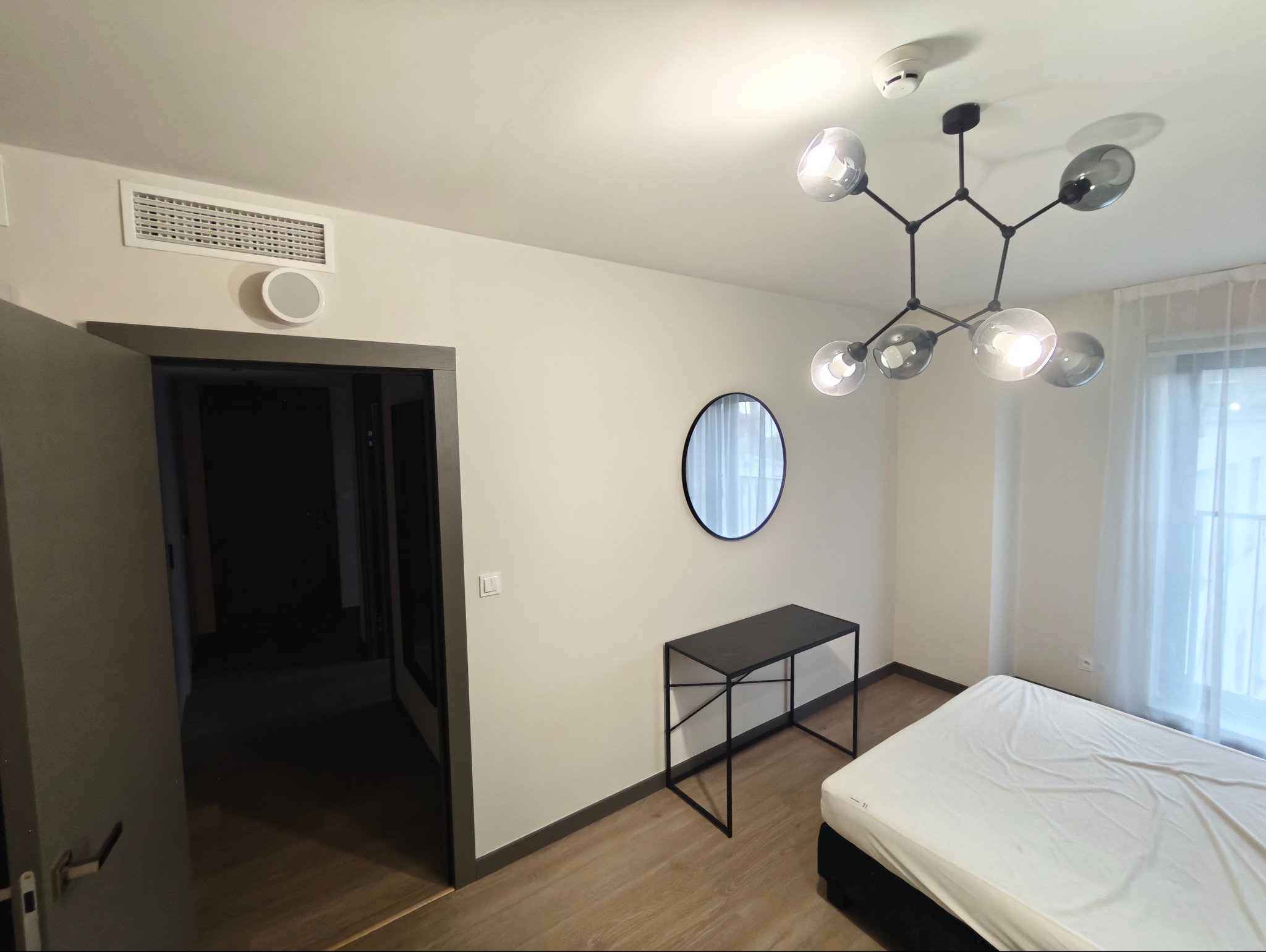 Jasne wnętrze pokoju z łóżkiem, czarną konsolą i okrągłym lustrem. Na suficie designerska lampa. Widoczny fragment wentylacji i drzwi do innego pomieszczenia.