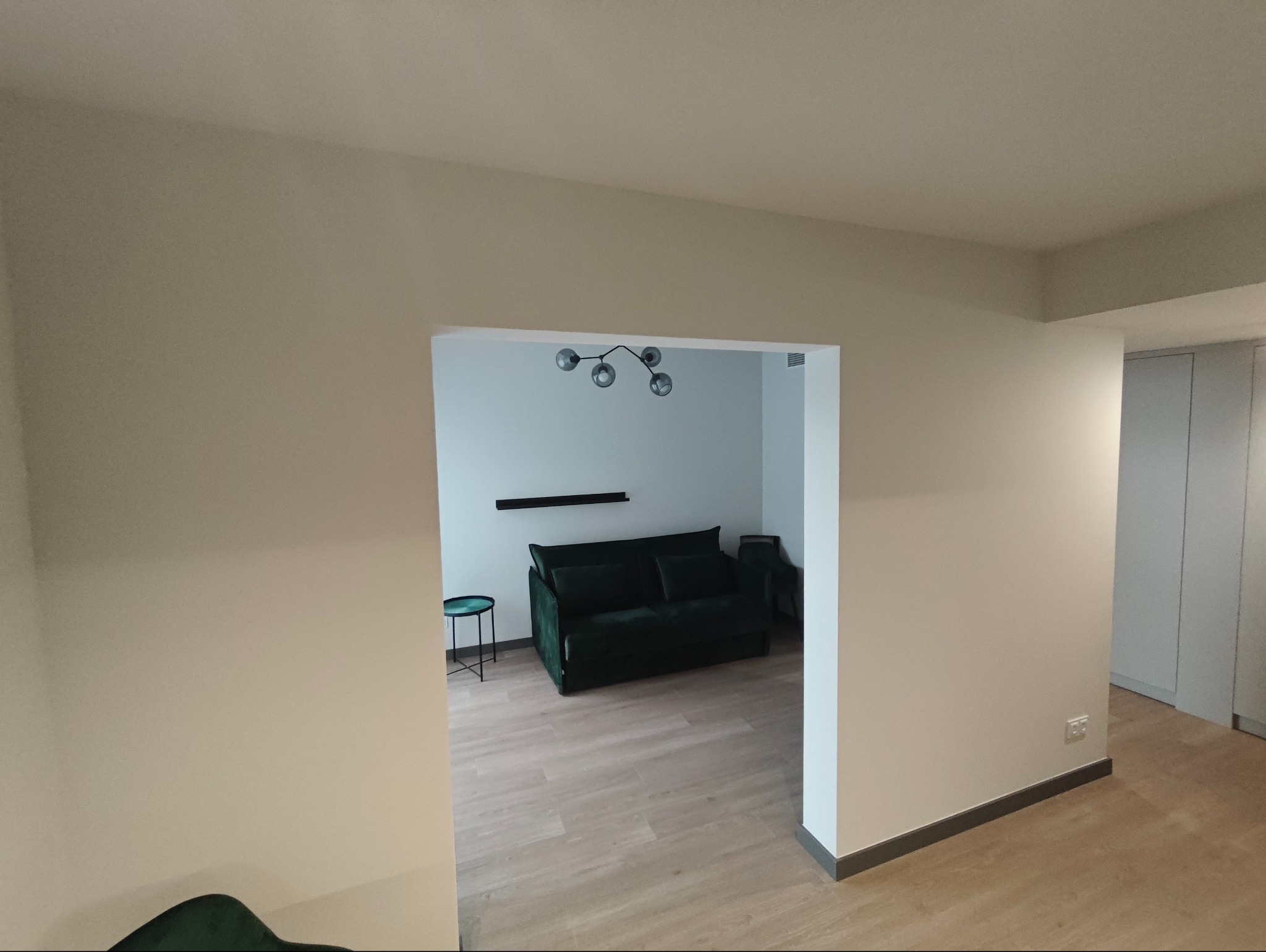 Jasne, świeżo pomalowane ściany w neutralnym odcieniu tworzą spokojne tło dla salonu z zieloną sofą. Widok na pokój z drewnianą podłogą i nowoczesnym oświetleniem.