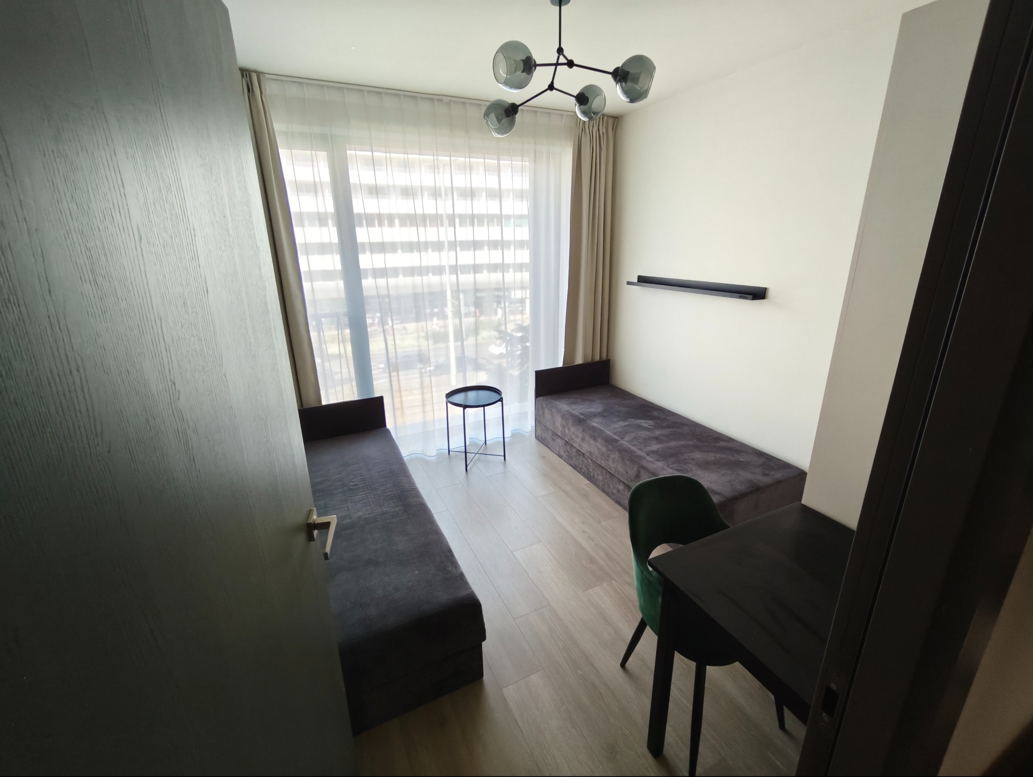 Nowoczesny pokój z dwoma szarymi tapczanami, stolikiem kawowym i designerską lampą sufitową. Białe ściany i jasne panele podłogowe. Widok zza otwartych drzwi.